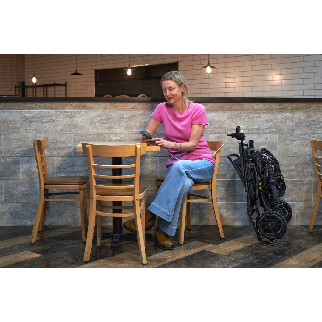 Golden Tech Cricket Carbon Fiber Foldable Travel Power Wheelchair - folded while user charges her phone